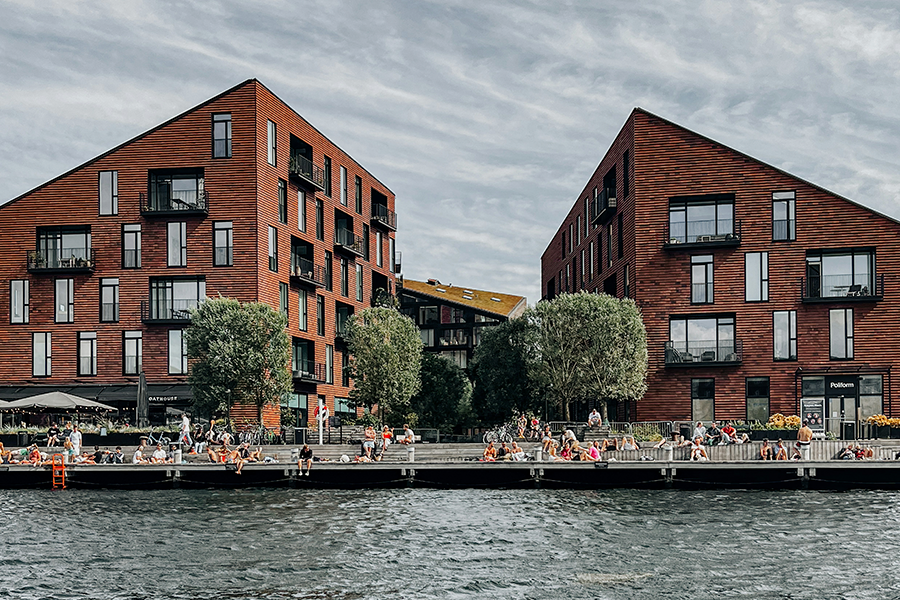 Du finder havnebade som dette på Christianshavn mange steder i København, hvor man ser to store bygninger nær havnebadet, hvor mange unge mennesker bader og slikker sol