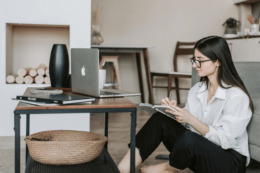 Kvindelig studerende  med langt mørkt hår, briller og hvid skjorte sidder på gulvet foran sin sofa med sin computer og søger efter en studiebolig